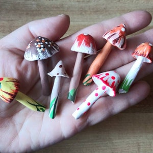 May include: A collection of eight hand-painted miniature mushroom figurines. The mushrooms are made of clay and feature various colors and patterns, including red, white, yellow, brown, and orange.