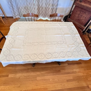 May include: A rectangular, off-white tablecloth with a detailed, openwork border. The border features a repeating pattern of interconnected ovals and lines. The tablecloth is spread over a wooden table, with two wooden chairs visible in the background.