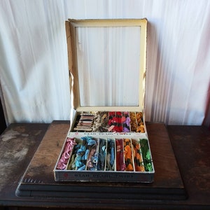 Puede incluir: Una caja de madera vintage llena de carretes de hilo de bordar de colores. La caja tiene una tapa de vidrio y está etiquetada "Star Pearl Twist" con un logotipo de estrella roja.