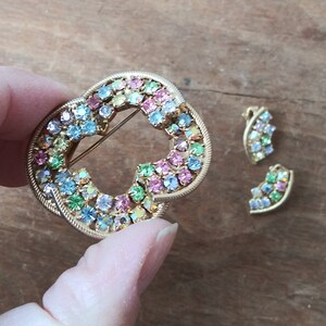 May include: A gold-toned brooch with a heart-shaped design, featuring a cluster of colorful rhinestones in shades of pink, blue, green, and yellow. Two smaller matching earrings are also included.