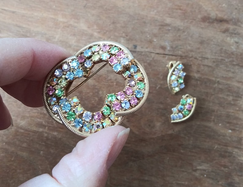 May include: A gold-toned brooch with a heart-shaped design, featuring a cluster of colorful rhinestones in shades of pink, blue, green, and yellow. Two smaller matching earrings are also included.