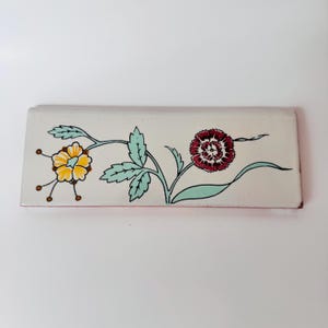 Peut inclure: Un carreau en céramique rectangulaire à fond blanc orné d'un motif floral peint à la main. L'illustration comprend une fleur jaune avec des tiges fines et une fleur rouge foncé avec des feuilles et des tiges vert clair sur la surface.