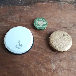 May include: Three round, vintage compact cases. One is white with a black rim and the text "Max Factor Hollywood U.S.A."  The second is green with a floral design. The third is gold with a floral design.