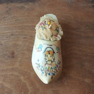 Op de afbeelding: Een houten naaldenkussen in de vorm van een klomp met een bloemenmotief en een geschilderd meisjesgezicht. Het naaldenkussen heeft een stoffen bovenkant met kant en is gevuld met naalden.