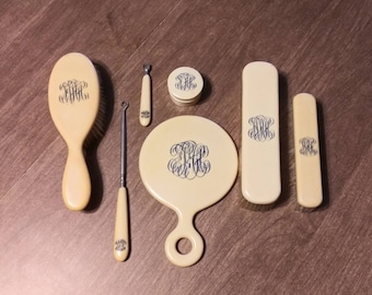 Può includere: Un set da toeletta antico con spazzola, specchio e altri accessori. Il set è color crema con un monogramma blu scuro. La spazzola è lunga circa 18 cm. Lo specchio è rotondo con un manico. Include un piccolo barattolo e uno strumento per unghie.
