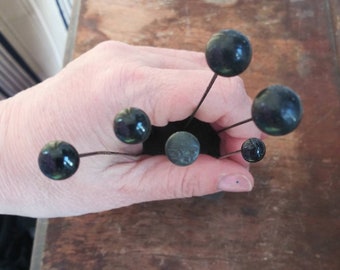May include: A set of six black glass-headed pins with metal shafts. The pins are arranged in a starburst pattern.