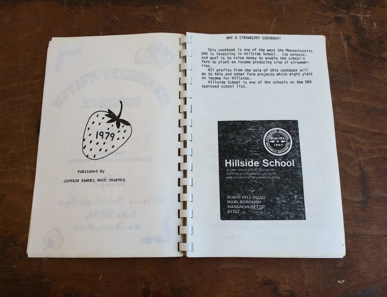 May include: Open cookbook titled "Strawberry Kisses Cookbook 1979" with a strawberry illustration. The book is published by Captain Samuel Wood Chapter. The right page has text about the cookbook's purpose and a Hillside School logo.