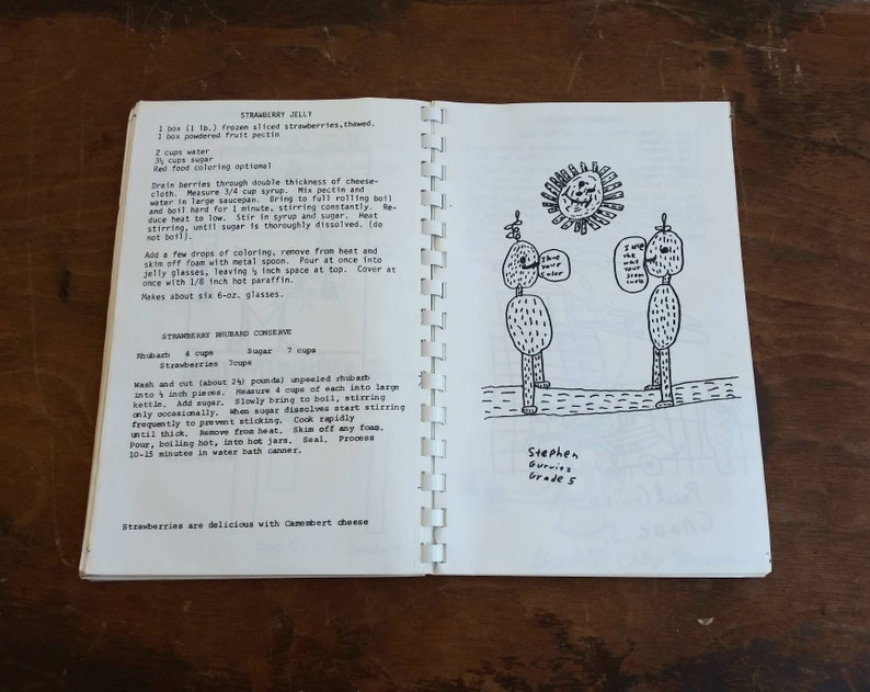 May include: Open cookbook pages with handwritten recipes for strawberry jelly and rhubarb conserve. A whimsical drawing of two figures with speech bubbles and a sun is on the right page. The text reads "I like your color" and "I wish the world was..."