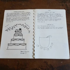 May include: Open spiral-bound book with handwritten recipes for champagne wedding punch, champagne frost punch, and champagne cup. The left page features a drawing of a tiered wedding cake. The book is open on a wooden surface.