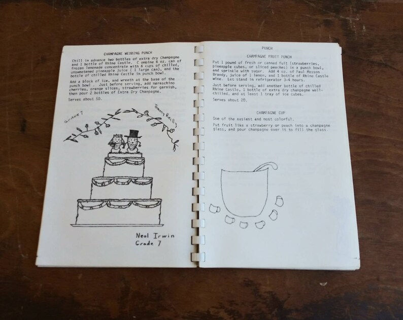 May include: Open spiral-bound book with handwritten recipes for champagne wedding punch, champagne frost punch, and champagne cup. The left page features a drawing of a tiered wedding cake. The book is open on a wooden surface.