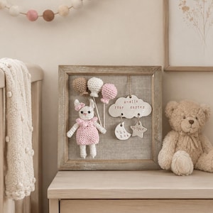 Puede incluir: Decoración enmarcada para guardería con un conejito de ganchillo sosteniendo adornos en forma de globo. El marco incluye una etiqueta en forma de nube con los nombres "Amelia Rose Carter" y detalles del nacimiento. También se ve un osito de peluche y una guirnalda.