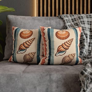 May include: Two square cushions with a cream background and vertical stripes in blue, coral, and white. Each cushion features illustrations of seashells in shades of brown and orange, creating a coastal theme.