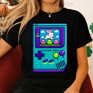 以下が含まれることがあります： 黒いTシャツ。ピクセル化されたグラフィックで、ウサギとイースターエッグが描かれた携帯ゲーム機が特徴です。「Easter Mode」と表示されたゲーム画面。コンソールはティールと紫で、緑と紫のボタンが付いています。