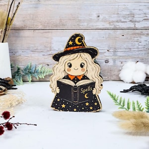 May include: A wooden cutout decoration featuring a cartoon character with long wavy hair wearing a black witch hat with a yellow crescent moon and star. The character holds an open black book with the word Spells and a crescent moon printed on it.