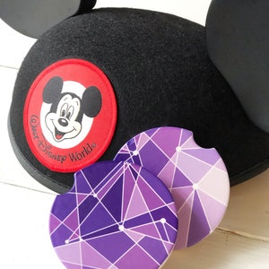 Puede incluir: Orejas de Mickey Mouse negras con un parche rojo que presenta la cara de Mickey Mouse y el texto "Walt Disney World". Dos posavasos geom&eacute;tricos morados y blancos con fondo blanco.