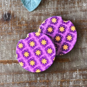 May include: Two purple and yellow round drink coasters with a repeating pattern of a sun and swirls.