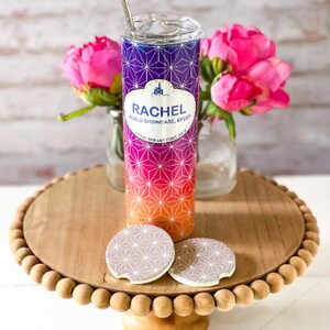 May include: A stainless steel tumbler with a pink, orange, and purple geometric pattern. The tumbler has a white band with the text "Rachel World Showcase Epcot" and "Where Dreams Come True". The tumbler is sitting on a wooden pedestal with two coaster-like circles.