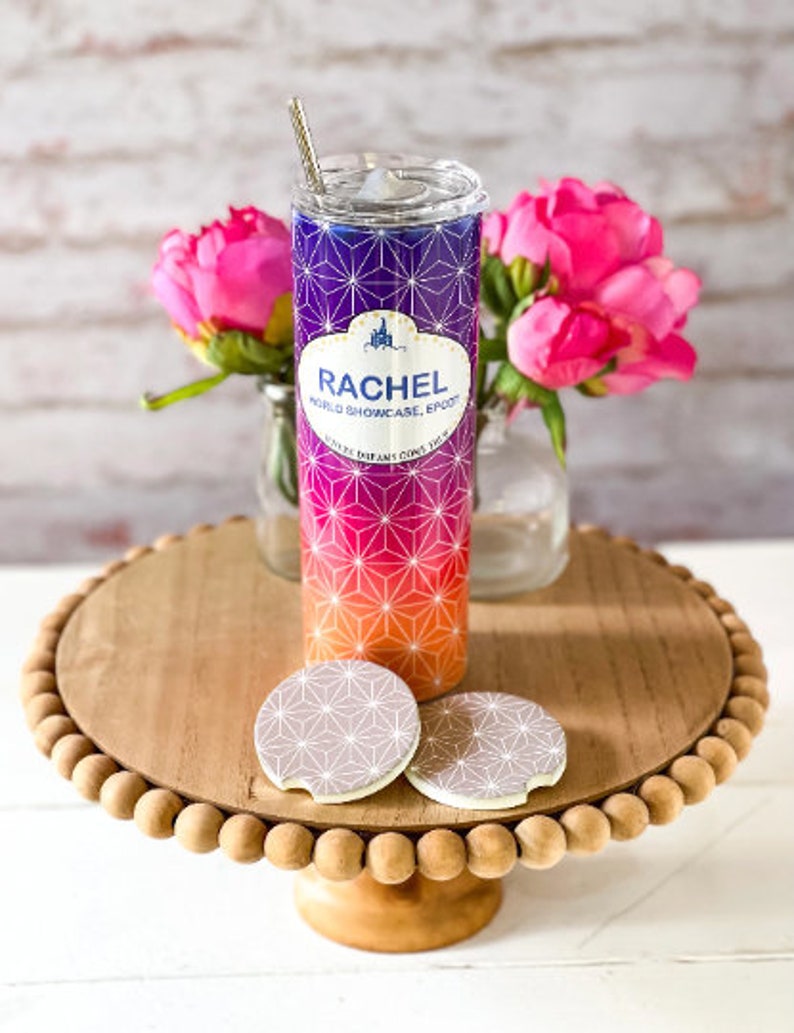 May include: A stainless steel tumbler with a pink, orange, and purple geometric pattern. The tumbler has a white band with the text "Rachel World Showcase Epcot" and "Where Dreams Come True". The tumbler is sitting on a wooden pedestal with two coaster-like circles.