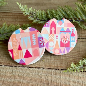 May include: Two pink and white ceramic coasters with a colorful geometric pattern inspired by a famous theme park. The coasters feature a whimsical design with a castle silhouette and numbers.