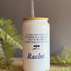 May include: White ceramic tumbler with a bamboo lid and a clear drinking straw. The tumbler has a quote about dragons and riders, and the name "Rachel" is printed on the bottom.