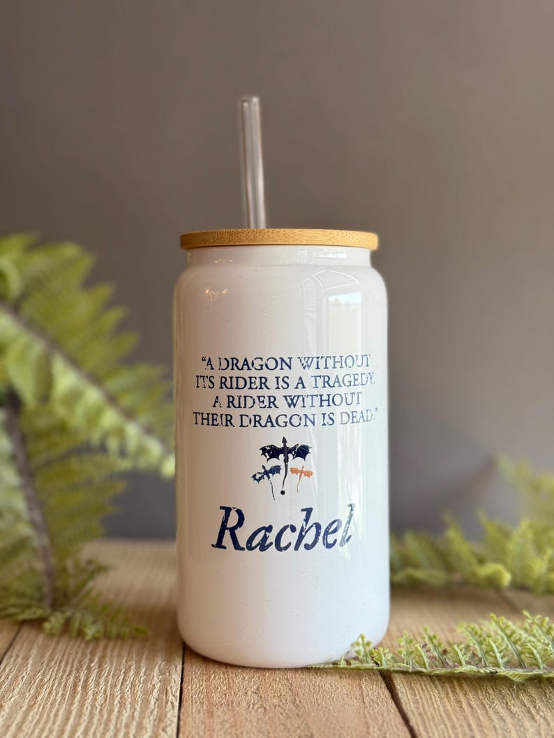 May include: White ceramic tumbler with a bamboo lid and a clear drinking straw. The tumbler has a quote about dragons and riders, and the name "Rachel" is printed on the bottom.