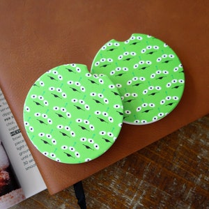 May include: Two green and white round coasters with a repeating pattern of alien eyes. The coasters are made of ceramic and have a cork backing.