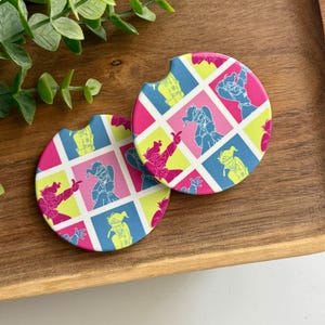 May include: Two circular car coasters featuring a grid pattern of colorful squares in shades of bright pink, lime green, and blue. Each square contains a stylized line art illustration of a character in various poses, with a small notch on the edge.