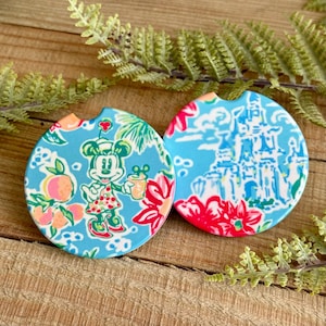 May include: Two round blue and white patterned coasters. One coaster features a cartoon image of Minnie Mouse in a tropical setting with pink and orange fruit. The other coaster features a white castle with blue and pink floral accents.
