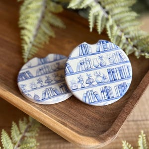 May include: Two round car coasters with a blue and white bookshelf design. The coasters feature a repeating pattern of books and vases. They are displayed on a wooden tray with green foliage.