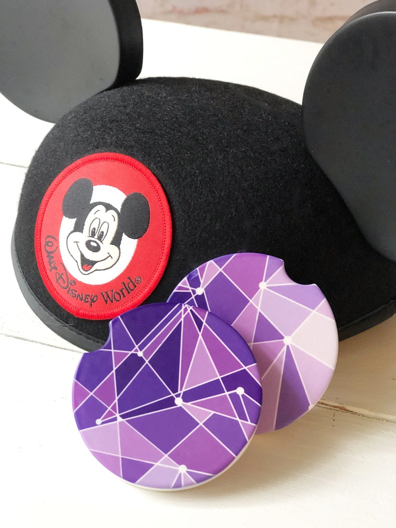 Puede incluir: Orejas de Mickey Mouse negras con un parche rojo que presenta la cara de Mickey Mouse y el texto "Walt Disney World". Dos posavasos geom&eacute;tricos morados y blancos con fondo blanco.