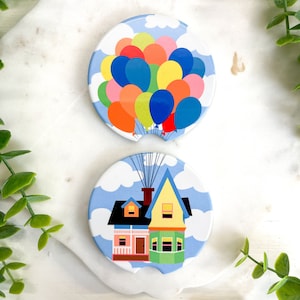 May include: Two round ceramic coasters with a blue sky and white clouds background. One coaster features a house with a yellow roof and green trim, while the other coaster features a bunch of colorful balloons.