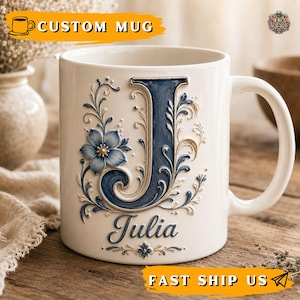 Pode incluir: Uma caneca de cerâmica branca com uma letra J grande em relevo na cor azul-escura, decorada com volutas florais intrincadas e uma flor azul. O nome Julia está escrito em uma elegante letra cursiva azul-escura abaixo da letra principal.