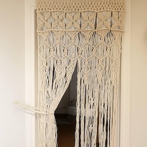 May include: Cream-coloured macrame door curtain. The curtain has a geometric design at the top and long, vertical strands that hang down. The curtain is tied back to reveal the doorway. The macrame is made of a thick, woven material.