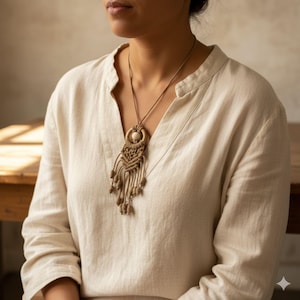 May include: A beige macrame necklace with a circular pendant and dangling tassels. The necklace is worn over a cream-coloured linen shirt. The pendant features a light-coloured bead in the centre.