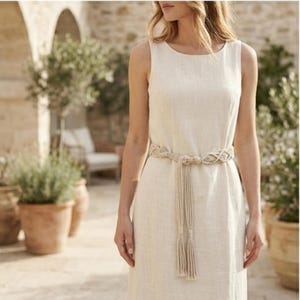 May include: A cream-coloured, sleeveless linen dress with a macramé belt. The belt is knotted at the waist and has a tassel detail. The dress has a simple, elegant design, suitable for warm weather.