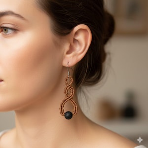 Może przedstawiać: Długie, brązowe kolczyki z makramy z czarną koralikiem. Kolczyki mają pleciony wzór i zwisają. Wykonane są z jasnobrązowego materiału i mają srebrne haczyki.