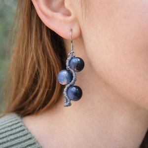 May include: Handmade earrings with three dark blue spherical beads. The beads are linked by a grey, woven cord in a spiral pattern. The earrings hang from silver-coloured hooks, offering a unique, artistic style. Each bead is approximately 1.5 cm in diameter.