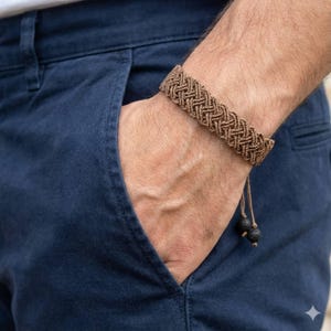 Op de afbeelding: Een bruine macramé armband met een gevlochten patroon. De armband wordt om de pols gedragen en heeft een trekkoordsluiting met twee kleine zwarte kralen. De persoon draagt een marineblauwe broek.