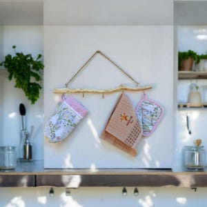 May include: A rustic wooden branch with hooks hangs on a white wall. It is decorated with two pink and white floral oven mitts and a brown waffle weave kitchen towel. The towel has the text "Made with Love" embroidered on it.