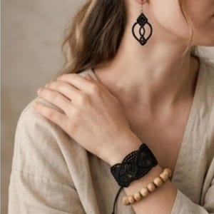May include: Black lace earrings and bracelet set. The earrings have a geometric design. The bracelet is made of black lace and a wooden bead bracelet. The jewelry is worn on a person's arm and ear.
