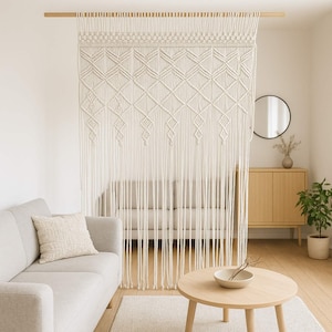 Puede incluir: Cortina de macramé color crema con un patrón geométrico de diamantes, suspendida de una barra de madera. La cortina cuelga frente a un sofá, creando un separador de ambientes suave y texturizado. El diseño presenta intrincados nudos y flecos largos y fluidos.