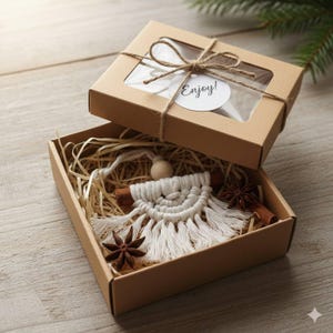 May include: A brown gift box with a clear window, tied with twine, and a tag saying "Enjoy!". Inside, a macrame ornament, cinnamon sticks, star anise, and straw. The box is on a wooden surface, with a sprig of fir in the background.