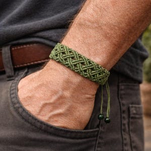 May include: A green macrame bracelet with a woven design. The bracelet is on a wrist, and the person is wearing a dark gray shirt and pants. The bracelet has adjustable green cords and two green beads.