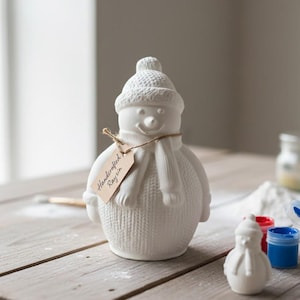Könnte beinhalten: Eine weiße Schneemannfigur aus Keramik mit gestrickter Mütze, Schal und Körper. Ein kleines Etikett trägt die Aufschrift "Handcrafted Raysin". Eine kleinere Schneemannfigur und Farbtöpfe befinden sich in der Nähe. Der Schneemann steht auf einer Holzoberfläche.