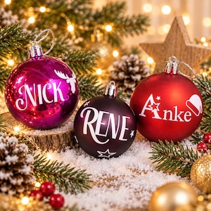 May include: Three round Christmas ornaments with silver hanging loops. The left ornament is shiny magenta with the white text "NICK" and a reindeer silhouette. The center is matte black with the white text "RENE" and stars. The right is matte red with the white text "Anke" and a Santa hat.