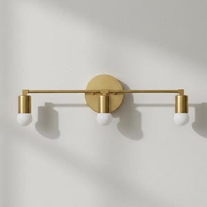 May include: A gold-toned, three-light vanity fixture. The fixture has a circular base and a horizontal bar with three cylindrical light holders. Each holder has a white, round bulb. The fixture is mounted on a white wall.
