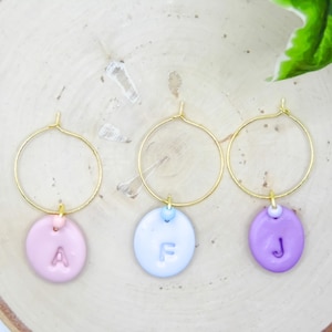 May include: Three gold-colored hoop earrings with pastel-colored charms. Each charm is a different color: pink, light blue, and purple. Each charm has a letter: A, F, and J. The earrings are displayed on a wooden surface.