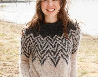 Pode incluir: Uma mulher vestindo um suéter de malha bege e cinza com um padrão de chevron preto. O suéter tem gola e punhos cinza escuro. A mulher está sorrindo e está ao ar livre perto da água.