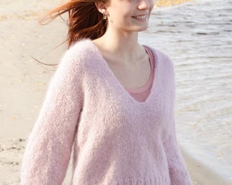 May include: A light pink, V-neck sweater with long sleeves. The sweater is made of a soft, textured material. The person is wearing a pink undershirt and white pants. The background is a beach with water and sand.