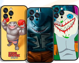 May include: Three phone cases with shark-themed designs. One case is yellow with a cartoon shark in boxing gloves, the text "KING SHARK Balboa". Another case features a blue shark reading a book. The third case has a shark with a colorful design.